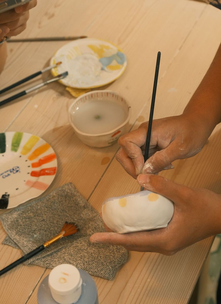 Mãos a pintar uma peça de cerâmica, com pincéis e tintas na mesa