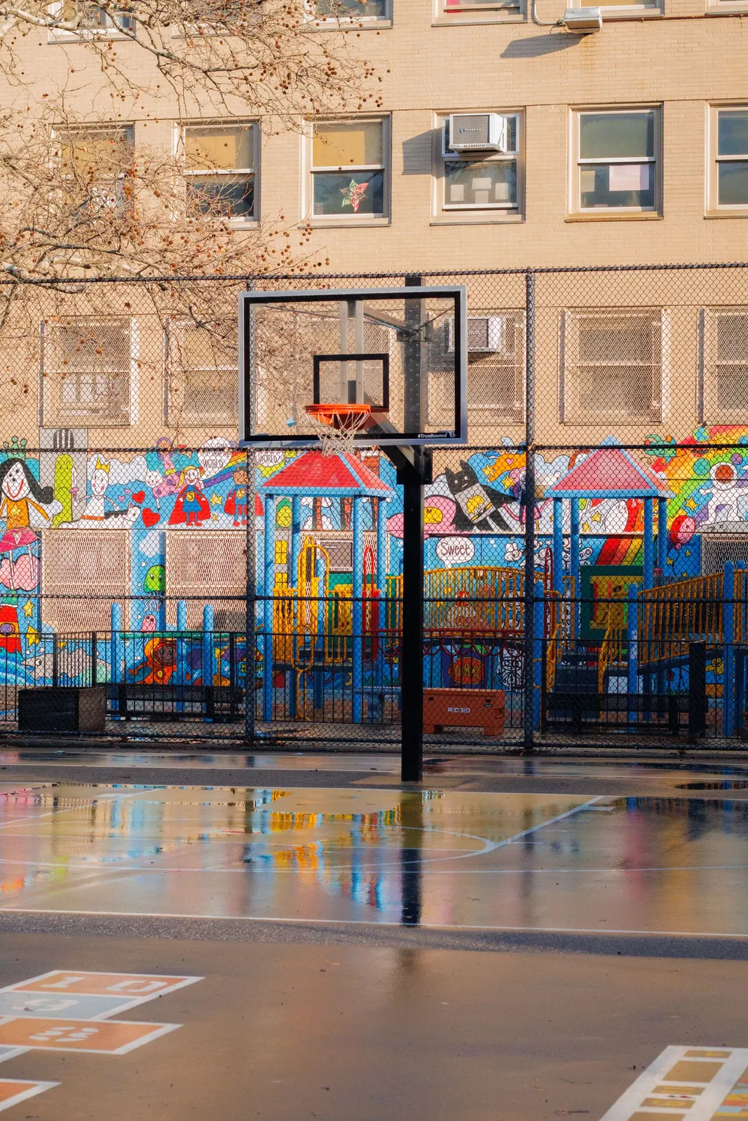 Pátio com tabela de basquetebol, parque infantil e um mural colorido por trás da rede