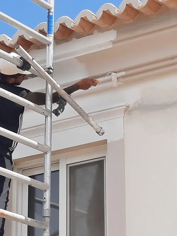 Pintor num andaime a pintar com rolo a cornija de uma fachada branca, junto ao beiral de telha