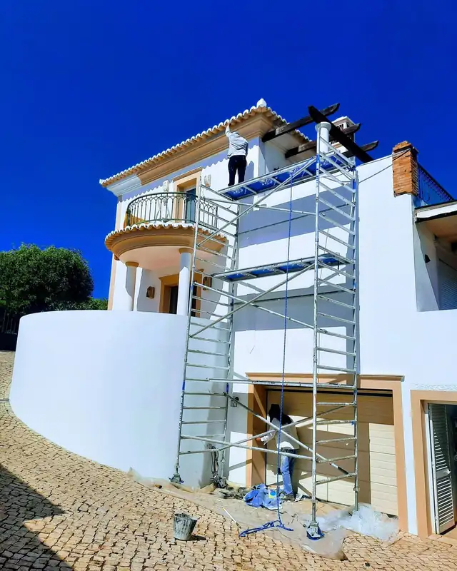 Técnicos a pintar a fachada de uma moradia com varanda redonda, com andaime montado e céu azul