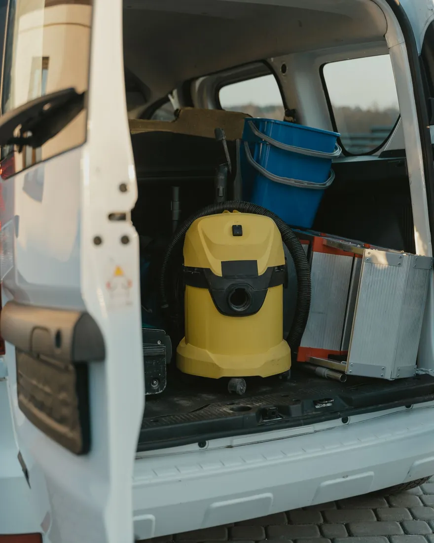 Carrinha branca de porta traseira aberta, com um aspirador de líquidos amarelo, um balde azul e uma escada lá dentro.